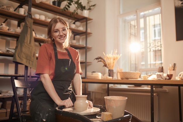 Warm getöntes Porträt einer jungen Handwerkerin, die in die Kamera lächelt, während sie in einer sonnenbeschienenen Werkstatt an der Töpferscheibe arbeitet und Kunst und Kunsthandwerk genießt, Platz kopieren