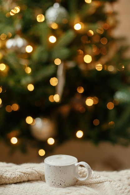 Warme Tasse Kaffee auf dem Hintergrund des Weihnachtsbaums mit Lichtern Gemütliches Zuhause Atmosphärische Winterhygge Stilvolle Tasse auf Strickpullover gegen goldene Beleuchtung Bokeh Weihnachtsbanner