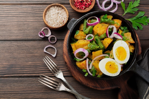 Warmer Kartoffelsalat mit grünen Bohnen, Pfeffer, Petersilie, Eiern und roten Zwiebeln in der Pfanne auf altem Holztisch. Selektiver Fokus. Draufsicht. Speicherplatz kopieren.