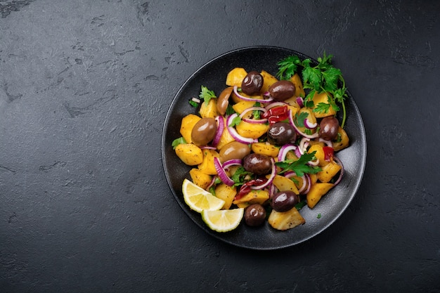 Warmer Kartoffelsalat mit Oliven, Pfeffer, Petersilie und roten Zwiebeln auf schwarzer Keramikplatte auf dunkler Betonoberfläche