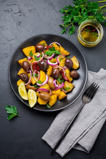 Warmer Kartoffelsalat mit Oliven, Pfeffer, Petersilie und roten Zwiebeln auf schwarzer Keramikplatte auf dunkler Betonoberfläche