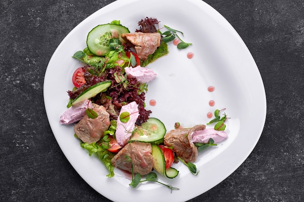 Warmer Salat mit Kalbfleisch und Frischkäse auf einem weißen Teller und einem dunklen Tisch