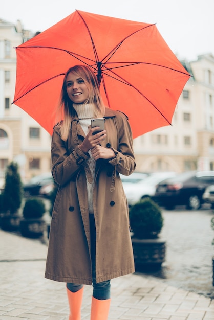 Warte auf Freund. Attraktive junge lächelnde Frau mit Regenschirm und Handy beim Stehen auf der Straße