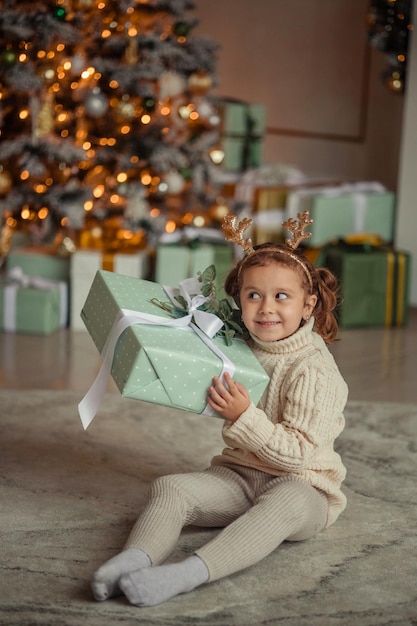 Warten auf das neue emotionale Porträt eines kleinen Mädchens zu Hause in einem Raum neben dem Weihnachtsbaum mit einem Geschenk in den Händen