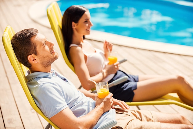 Was für ein großartiger Tag! Seitenansicht eines schönen jungen Paares, das Cocktails hält und lächelt, während es auf den Liegestühlen am Pool sitzt?