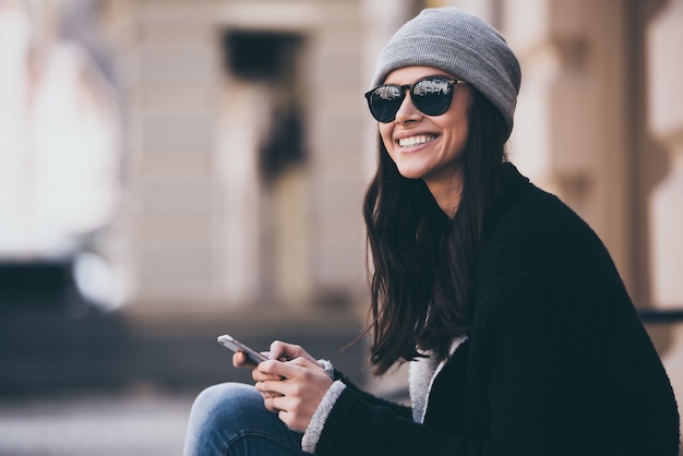 Was für ein schöner Tag! Seitenansicht einer schönen jungen Frau mit Sonnenbrille, die ihr Smartphone benutzt und mit einem Lächeln wegschaut, während sie im Freien sitzt
