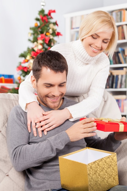 Was für ein schönes Geschenk! Schöner junger Mann, der auf der Couch sitzt und eine Geschenkbox hält, während ihre Freundin hinter ihm steht und lächelt