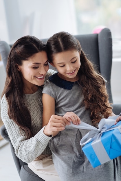 Was ist innen. Nettes angenehmes neugieriges Mädchen, das auf dem Schoß der Mutter sitzt und das Geschenk öffnet, während sie sich fragt, was drin ist