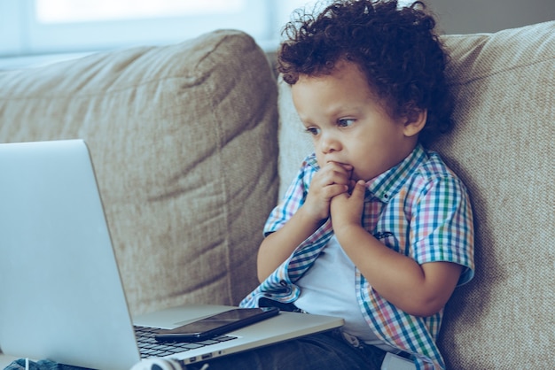 Was sonst zu tun? Kleiner afrikanischer Junge, der auf seinen Laptop schaut und sich die Nägel beißt, während er zu Hause auf der Couch sitzt