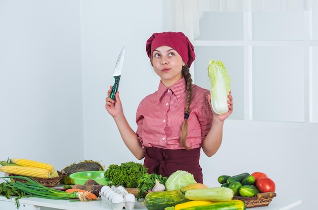 Was voller Vitamine zu kochen Kind trägt Kochuniform und Hut Teenager-Mädchen bereitet Chinakohl frisches rohes Gemüse zu Zeit, glückliches Kind zu essen, das in der Küche nur gesunde und biologische Lebensmittel kocht