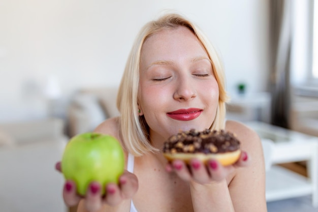 Was zu wählen gesund oder ungesund Porträt einer schönen jungen Frau, die zwischen einem Apfel und einem Donut wählt