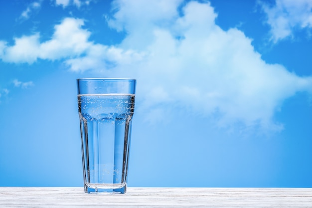 Wasser in großer Glasschale auf weißer Holzoberfläche. Blauer Hintergrund mit Wolken, Kopierraum.