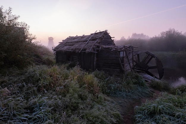 Wasser- und Windmühlenholzmühle am Fluss frühmorgens bei rosa Sonnenaufgang in einem traditionellen russischen Dorf. Ländliche Herbstlandschaft mit dem ersten Frost