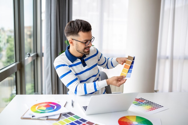 Web designer trabalhando em casa sentado à mesa com laptop moderno olhando amostras de cores homem feliz freelancer aproveitando seu espaço de cópia de trabalho remoto