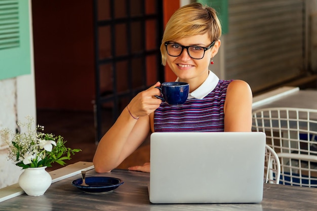 Webdesigner-FernarbeitskonzeptErfolgreiche stilvolle Touristin, die an einem modernen Laptop-Computer arbeitet und an einem Tisch im Café-Restaurant am Meer sitztfreiberufliche freiberufliche Fernarbeit