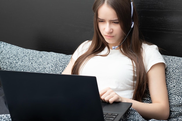 Webinar de educação on-line Um aluno em casa está envolvido no treinamento ouve atentamente e assiste a uma palestra em um laptop com um microfone em fones de ouvido deitado na cama