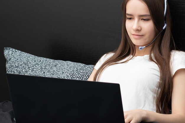 Webinar Girl en auriculares con un micrófono frente a una computadora portátil yace en la cama