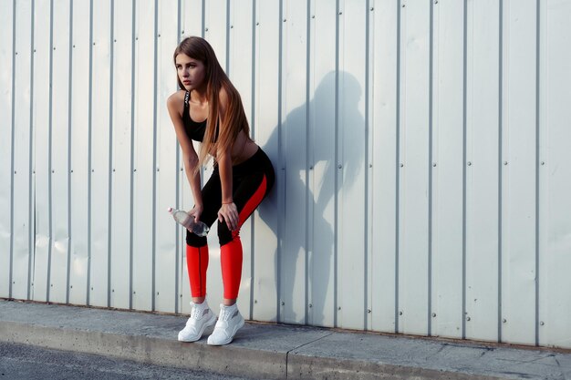 Foto weibliche athletin trinkt wasser, während sie an einer grauen wand steht, schlanke frau in sportbekleidung macht eine pause nach dem training