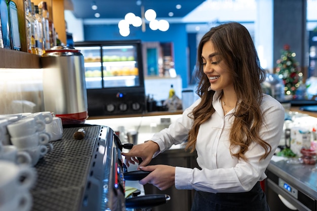 Weibliche Barista macht Kaffee an der Theke eines Cafés, Essen zum Mitnehmen, lächelnde Frau mit Schürze bereitet Kaffee für den Kunden vor