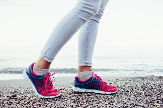 Weibliche Beine in rosa und blauen Turnschuhen und Jeans sind am Strand