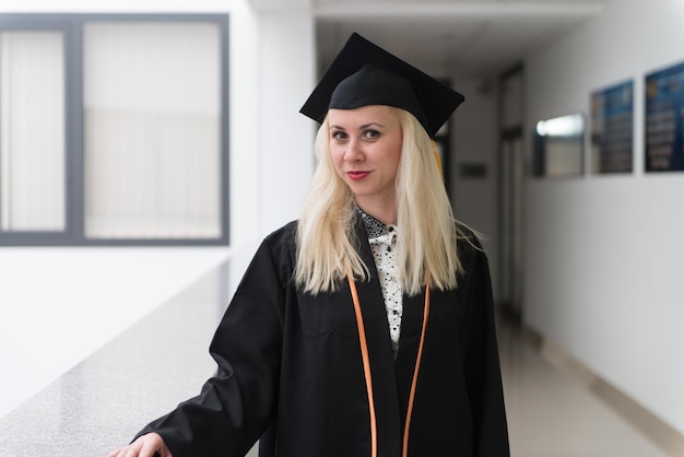 Foto weibliche blonde hochschul- oder gymnasiastin, die beim abschluss selbstbewusst schwarze mütze und kleid trägt