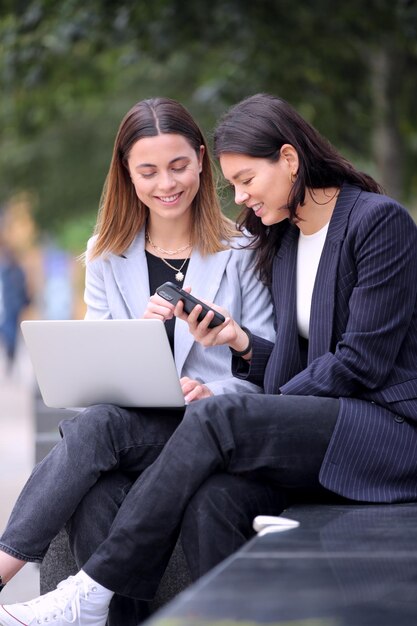 Weibliche Büroangestellte im Freien, die während der Büropause am Laptop arbeiten und mit dem Handy telefonieren