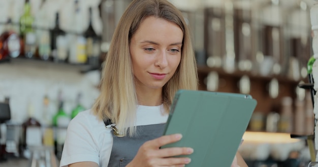 Weibliche Cafébesitzerin, die vor der Theke steht und einen digitalen Tablet-Computer in einer modernen Cafeteria verwendet. Frauenkellnerin oder Barista-Angestellter des Coffeeshops, der modernes Gerät hält.