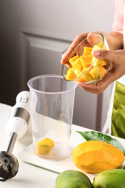 Weibliche Chefhand gießen gewürfelte Mango in den Entsafter-Bowl-Prozess, der Mango-Smoothie oder Mango-Lassi herstellt