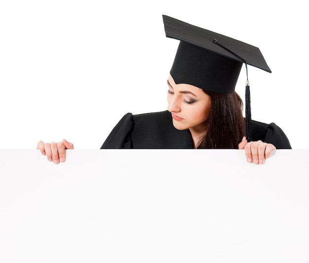 Weibliche Doktorandin späht hinter einer leeren Tafel hervor, isoliert auf weißem Hintergrund. Schöne Teenager-Studentin im Absolventenmantel, die ein leeres Plakat zeigt