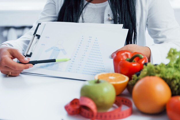 Weibliche Ernährungswissenschaftlerin in weißem Mantel sitzt im Innenraum im Büro am Arbeitsplatz und zeigt Informationen auf einem Dokument