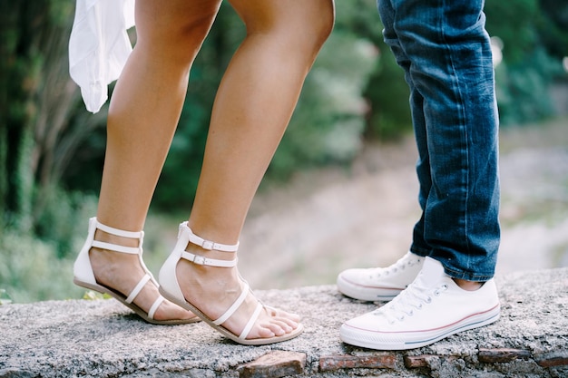 Weibliche Füße in Sandalen neben männlichen Füßen in Turnschuhen stehen auf einer Steingrenze im Park