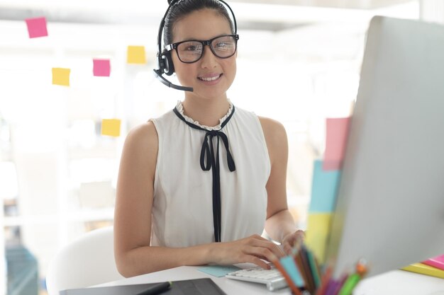 Weibliche Grafikdesignerin mit Headset, die in einem modernen Büro am Schreibtisch an die Kamera schaut