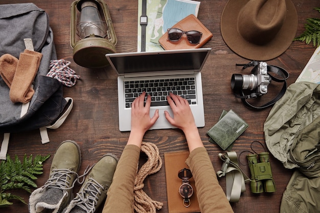 Weibliche Hände, die auf Laptop am Tisch mit wandernden Sachen wie Kleidung, Seil, Laterne, Karten tippen