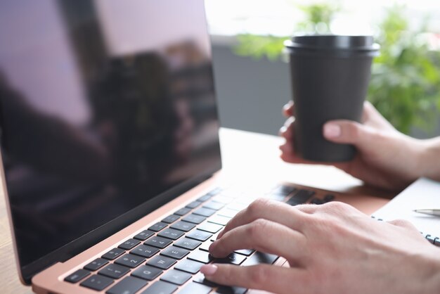 Weibliche Hände, die auf Laptop-Tastatur tippen, hält Glas mit Kaffee