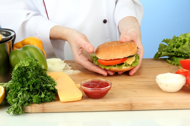 Weibliche Hände, die Cheeseburger auf blauem Hintergrund vorbereiten