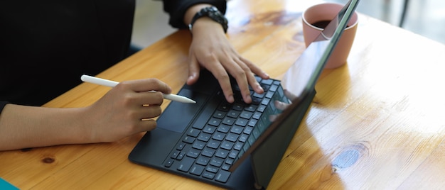 Weibliche Hände, die digitales Tablett mit Tastatur und Stift auf hölzernem Schreibtisch verwenden