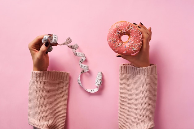 Weibliche Hände, die Donut halten und Band über rosa Hintergrund messen.