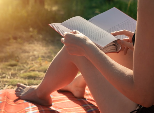 Weibliche Hände, die ein Buch zum Lesen von Sommerferienliteratur auf Plaid im Park in der Natur halten