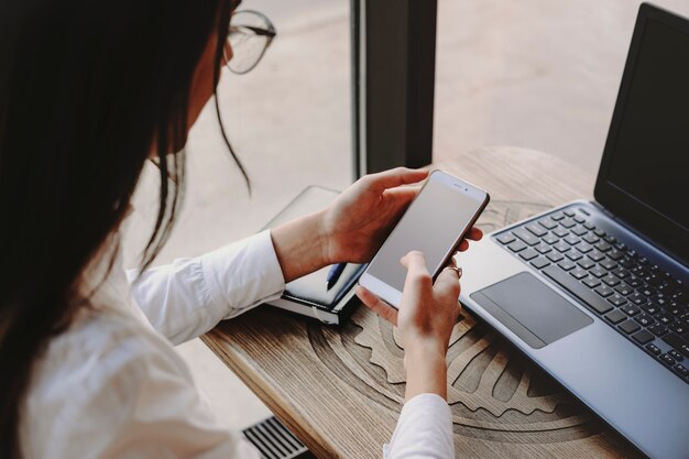 Weibliche Hände, die ein Smartphone für das Arbeiten an der Internet-Transaktion und dem Laptop verwenden, die an einem Tisch im Café sitzen.