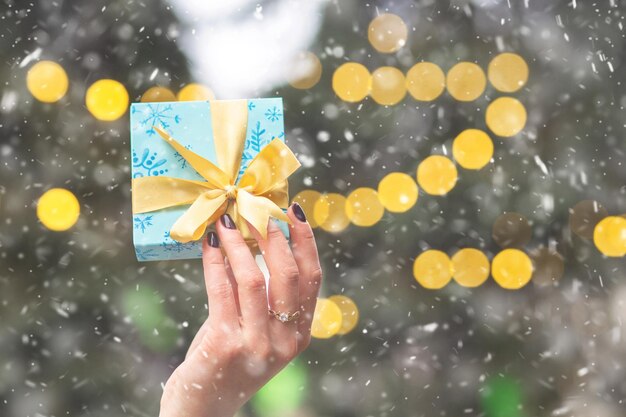 Weibliche Hände, die eine kleine Geschenkbox am Weihnachtsbaumhintergrund mit Bokeh-Lichtern halten. Freiraum