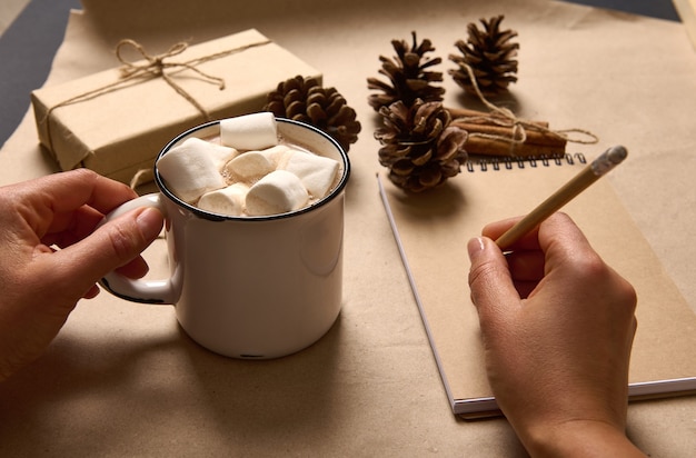 Weibliche Hände, die eine Tasse heißes Kakaogetränk und Marshmallows halten und mit einem Holzstift auf Kartonoberfläche mit Bastelweihnachtsgeschenk und Tannenzapfen auf ein leeres Notizbuchblatt schreiben