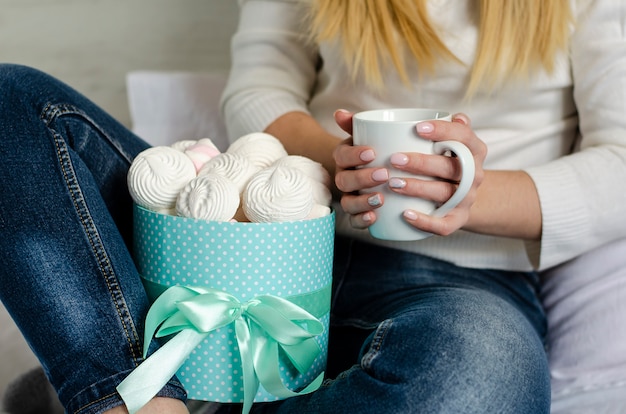 Weibliche Hände, die einen weißen Becher mit Lattekaffee halten. Marshmallows und Meringues in einer Box