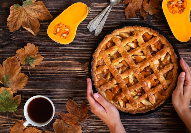 Weibliche Hände, die hausgemachten Kürbiskuchen halten. Halloween und Thanksgiving. Feiertagskürbisbonbons. Hölzerner Herbsthintergrund, trockene Blätter, geschnittener Kürbis, Tasse Tee. Draufsicht. Speicherplatz kopieren