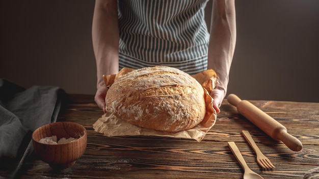 Weibliche Hände, die hausgemachtes Naturbrot mit einer goldenen Kruste auf einer Serviette auf einem alten Holzhintergrund halten Rustikaler Stil