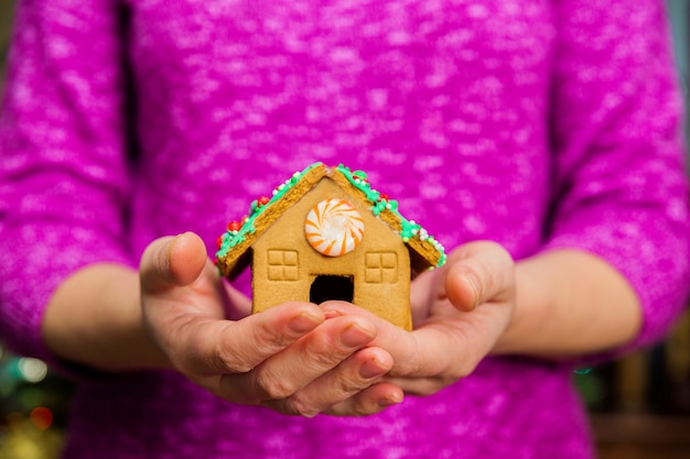 Weibliche Hände, die kleines Lebkuchenhaus auf Tisch mit Weihnachtsdekorationen halten