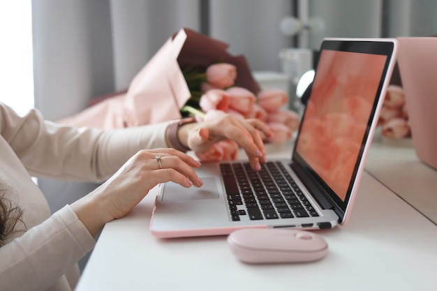 Weibliche Hände, die Laptop verwenden. Weiblicher Schreibtisch-Arbeitsplatz-Homeoffice-Mock-up mit Laptop, rosa Tulpenblumenstrauß, Smartphone und rosafarbenem Zubehör.