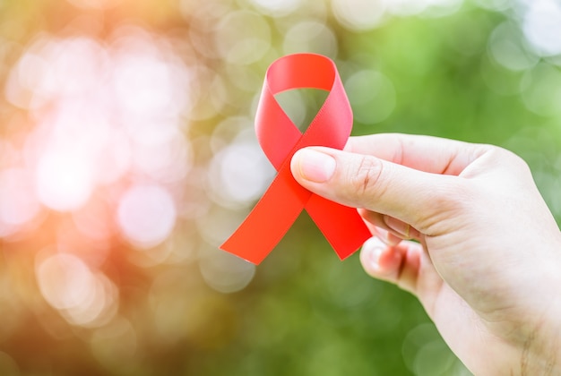 Weibliche Hände, die rotes AIDS-Bewusstseinsband auf grünem bokeh Hintergrund halten.