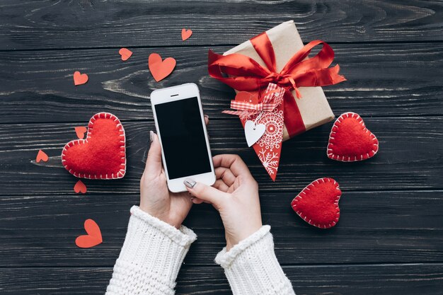Weibliche Hände, die Smartphone auf einem Hintergrund mit Geschenken, dekorative Herzen für Valentinstag halten.