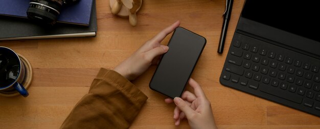 Weibliche Hände, die Smartphone auf Holztisch mit digitalem Tablett, Zubehör und Kaffeetasse halten