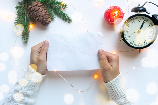 Weibliche Hände, gekleidet in einen weißen Pullover, halten ein Blatt handgemachtes weißes Papier. Leere Weihnachtsgrußkarte